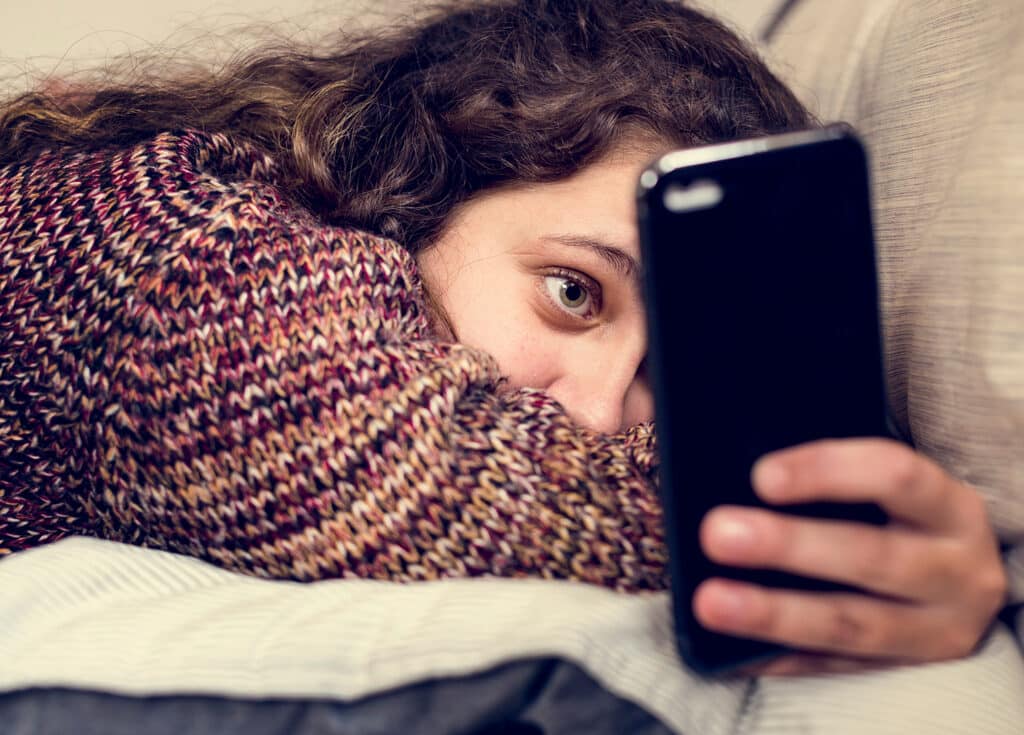 curly haired teen girl in sweater laying in bed looking at smartphone