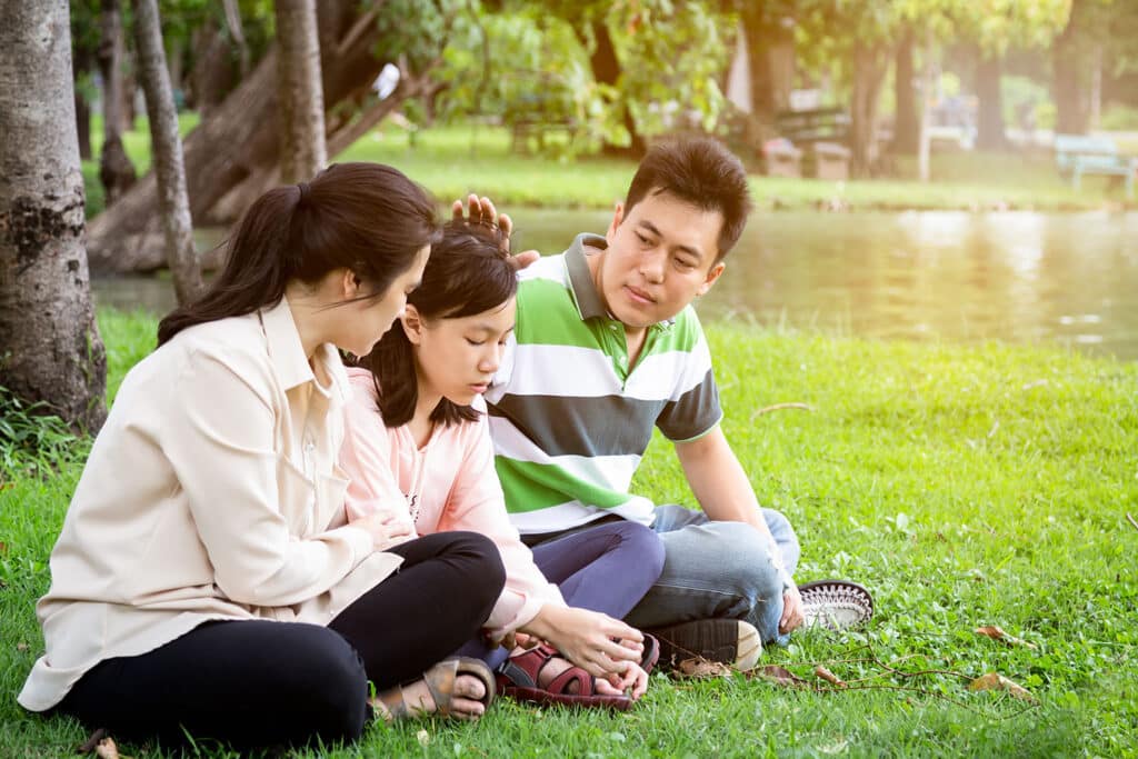 two parents sitting cross legged by their daughter by a lake