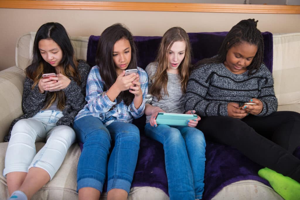 four young girls sitting next to each other on a couch looking at their own tech devices